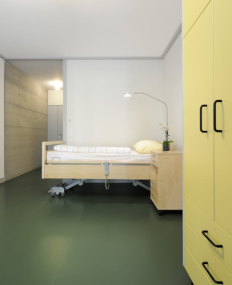 Bewohnerzimmer mit Pflegebett und passendem Nachttisch, Kleiderschrank mit gelben Türen rechts angeschnitten
