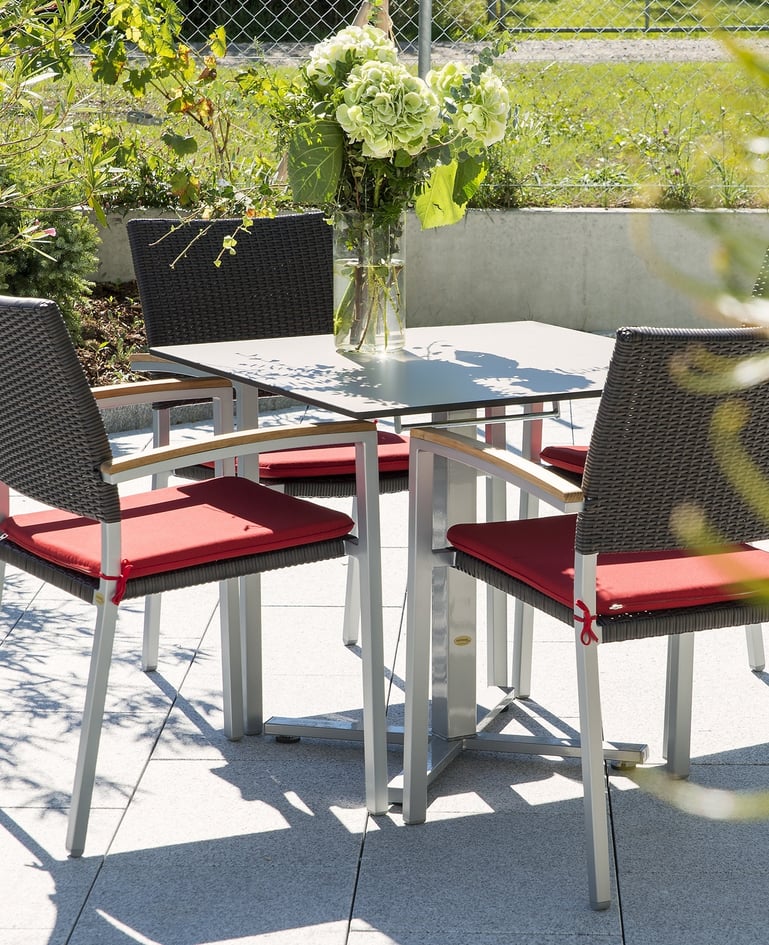 Vier Stühle mit Polster um Tisch mit Blumenvase gruppiert auf einer sonnigen Terrasse