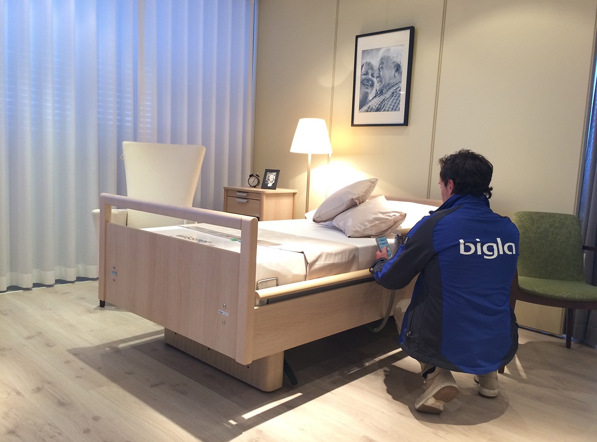 Fachperson von Bigla kniet an einem Pflegebett und nimmt mit der Handbedienung technische Einstellungen vor