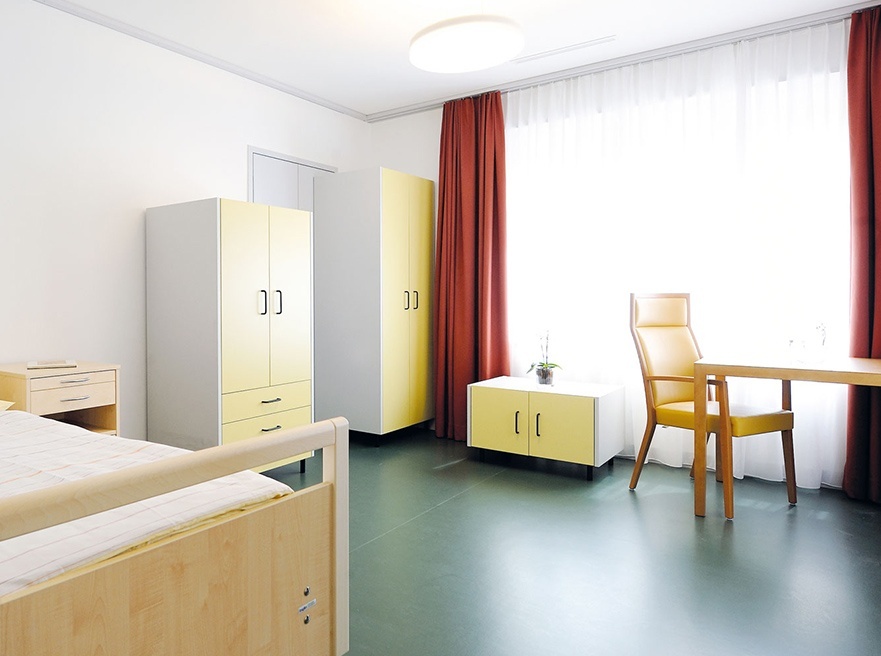 Unbewohntes Pflegezimmer, steril eingerichtet mit Linoleumboden, weissen Schrankmodulen und Pflegebett. Grosses Fenster und Tisch mit Stühlen