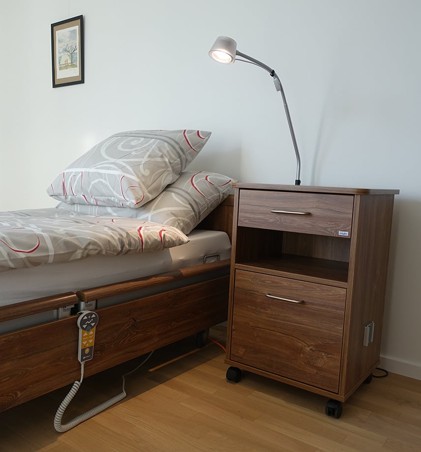 Pflegebett mit Handbedienung in Zimmer. Passender Nachttisch aus dunklem Holz mit Leselampe neben dem Bett