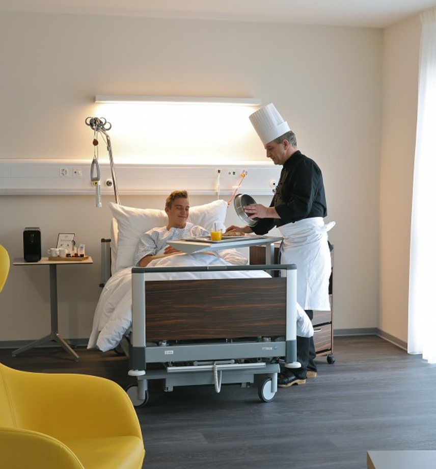 Koch serviert einem Patienten dessen Mahlzeit am Universalklinikbett