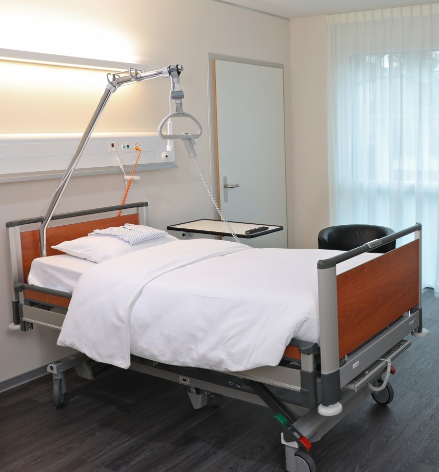 Spitalzimmer mit Universalklinikbett. Handbedienung am Triangelgriff eingehakt. Besuchersessel neben dem Bett